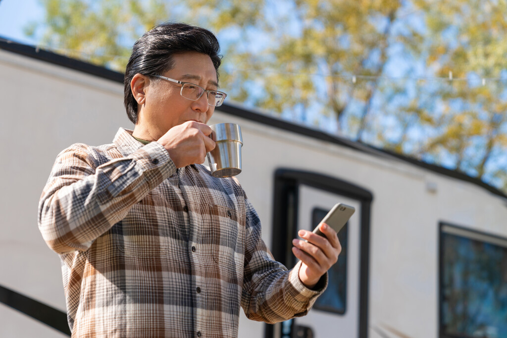 一个老年男人站在房车前看着手机视频喝咖啡图片